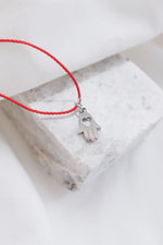 Silver hamsa hand pendant on red string necklace, symbolizing protection and blessings.